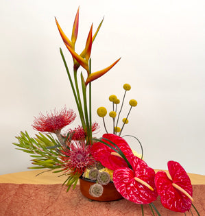 A striking tropical arrangement full of heliconia, pin cushion protea, anthurium, leucadendron, scabiosa pods, carnations, craspedia, and lily grass.