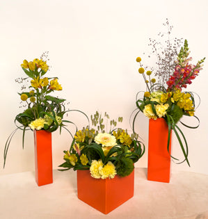 A combination of different vases with coordinating blooms in shades of yellow, such as, snapdragons, alstroemeria, carnations, dianthus, mini hydrangeas, craspedia, gerbera daises, and two types of quaking grasses, in armatures made of lily grass.