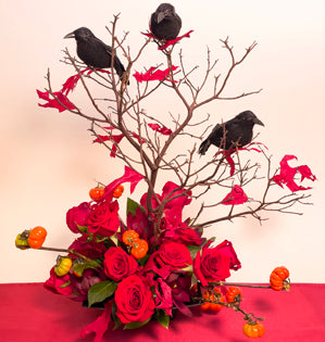 A bewitching Halloween design made with a variety of foliages, red roses, a manzanita branch, preserved leaves, ornamental pumpkins, and faux ravens.