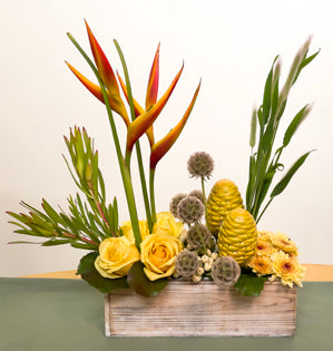 A charming arrangement in shades of yellow made with, parakeet heliconia, beehive ginger, chrysanthemums, roses, scabiosa, leucadendron, bunny tail grass, and galax leaves.