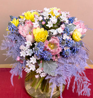 A petite hand-tied bouquet full of spring blooms such as, muscari, narcissus, allium, and fringed tulips, accented with dusty miller, and painted plumosa.