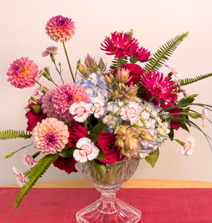 A glass compote full of summer blooms such as, hydrangea, dahlias, pasta gerberas, dianthus, roses, blushing bride protea, lily grass, and sword fern.