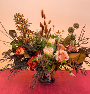 A lush compote design in shades of Mocha Mousse (The 2025 Pantone Color of the Year), full of magnolia leaves, nandina, agonis, ti leaves, foraged branches, scabiosa pods, moab and coffee break roses, miniature cymbidium orchids, protea, bunny tail grass, preserved beech leaves, clematis, pieris, and leucadendron. 